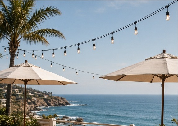 Shaded patio tables with umbrellas and string lights overlooking a blue cove