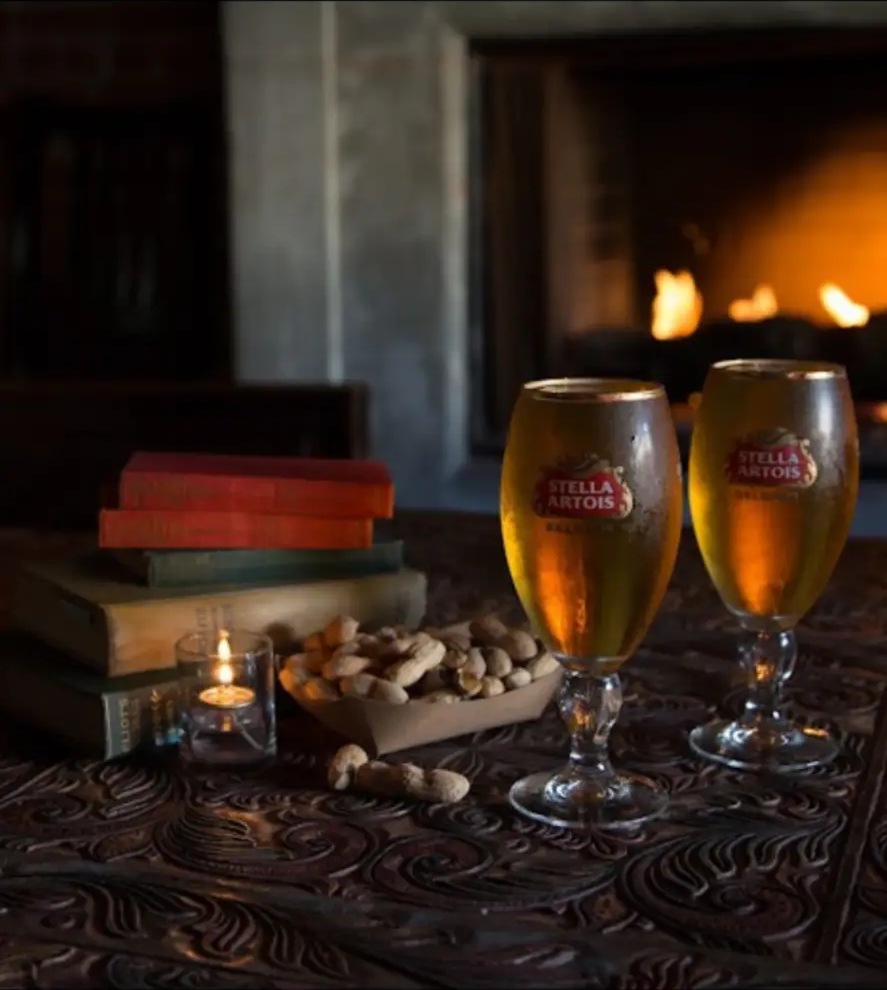 Two cold draft beers and a candle on a carved wooden table beside a lit fireplace
